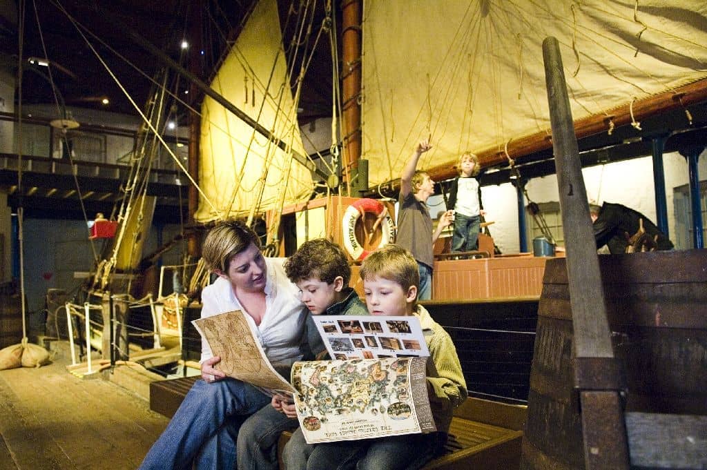 A woman and two children read museum maps inside the South Australian Maritime Museum, with a historic ship and sails in the background.