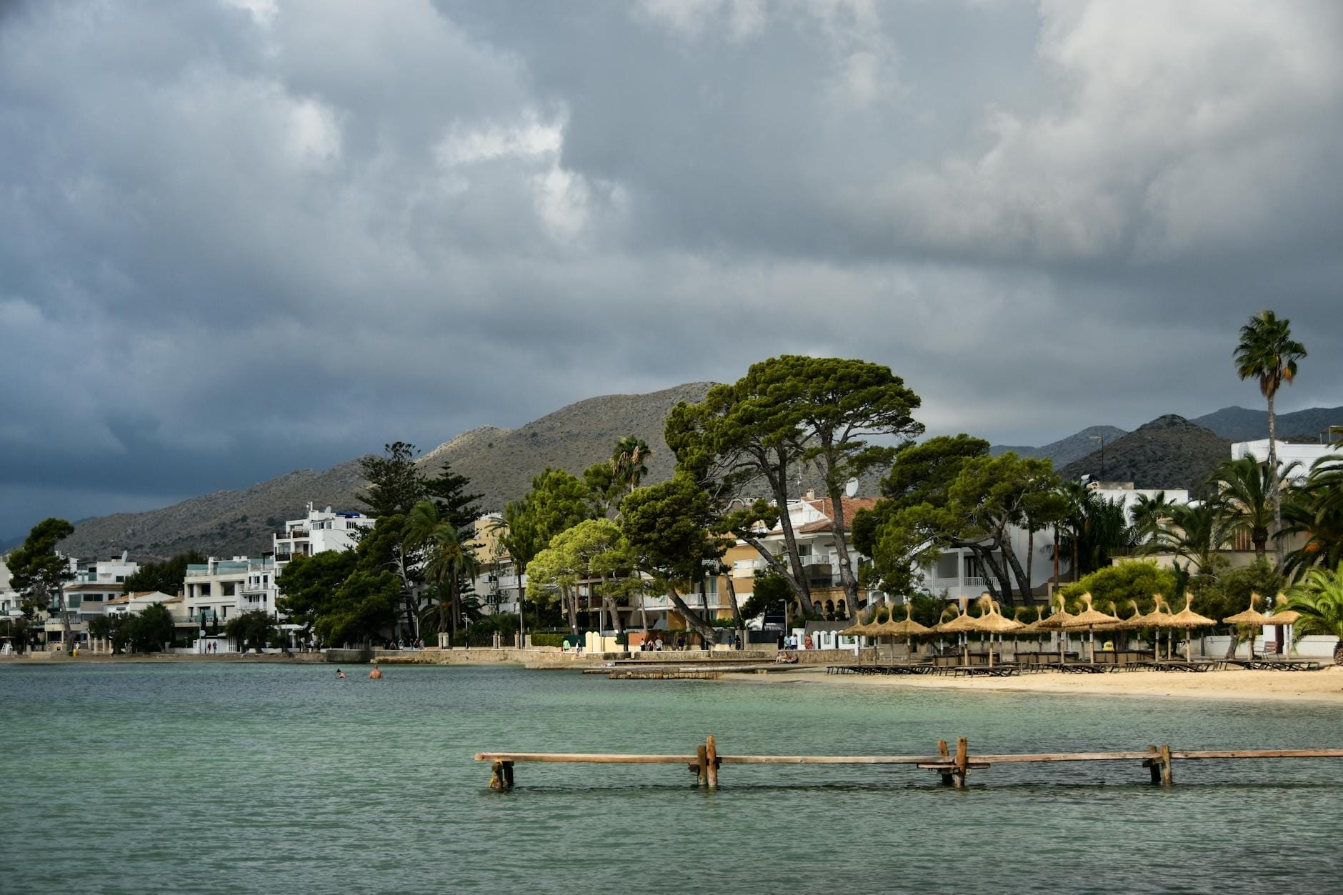 Una baia tranquilla con acqua turchese, pini, spiaggia sabbiosa e tradizionali edifici bianchi sullo sfondo delle montagne a Port de Pollença, Maiorca.