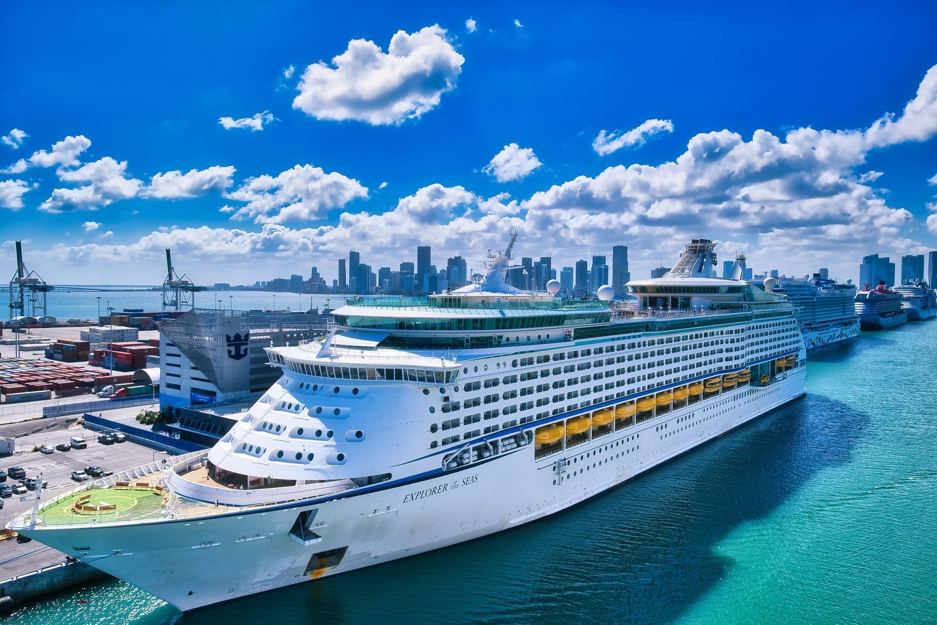 Grande nave da crociera ormeggiata al terminal di PortMiami con lo skyline del centro di Miami e un cielo azzurro sullo sfondo in una giornata di sole.