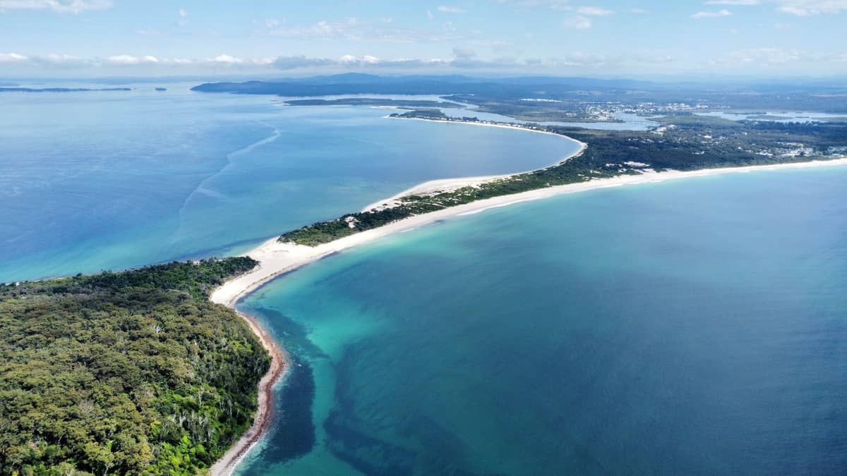Ein weitläufiger Luftblick auf Port Stephens mit langen Sandstränden, blaugrünem Wasser, Landzungen und bewaldeten Gebieten unter einem teils bewölkten Himmel.