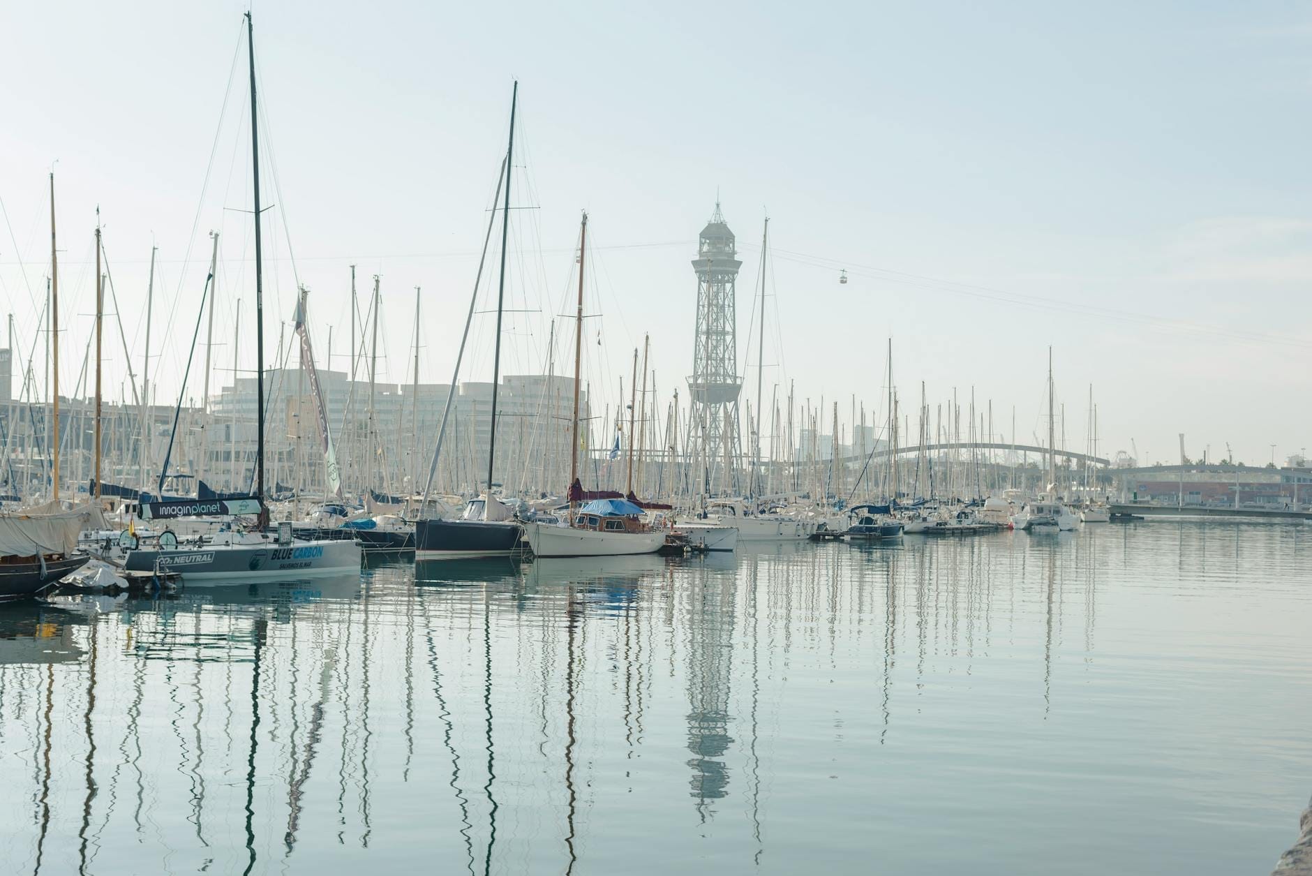 Wide view of Port Vell marina in Barcelona, with sailboats docked, calm water, and the iconic cable car tower visible in the background under a bright sky.