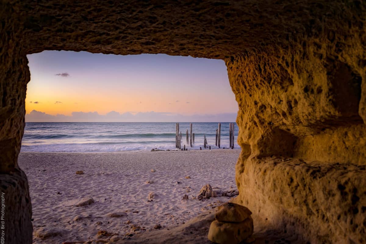 View of Port Willunga Beach through a red-ochre cave, featuring sandy shore, old jetty pylons, and calm sea at sunset.