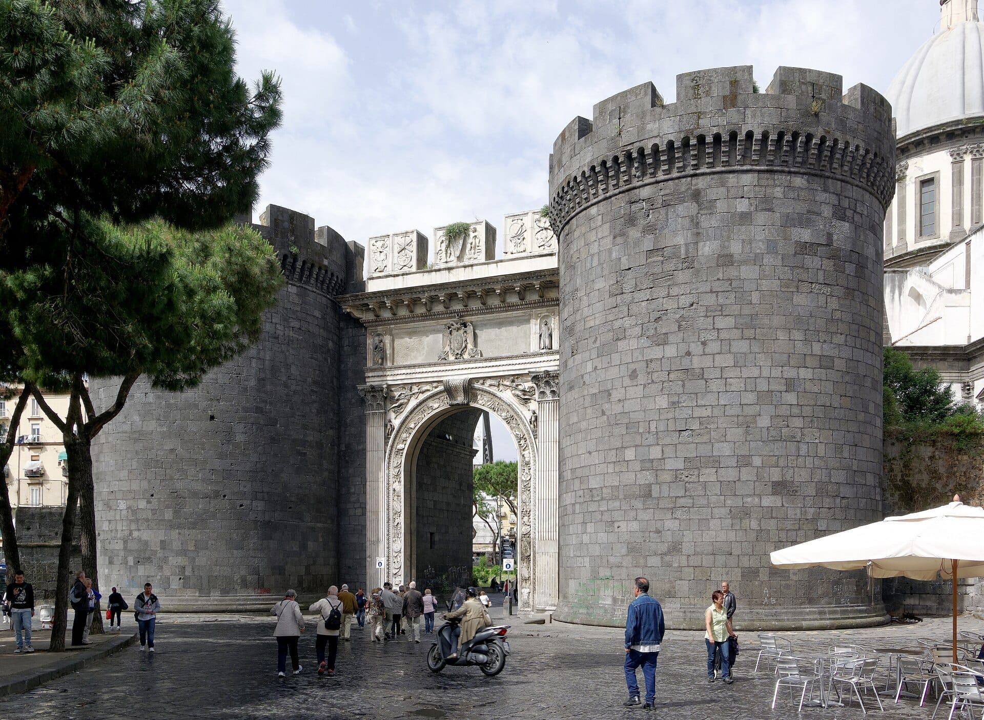 Porta Capuana a Napoli vista frontalmente, con le sue due imponenti torri in marmo, l'arco rinascimentale e alcune persone che camminano nei dintorni.
