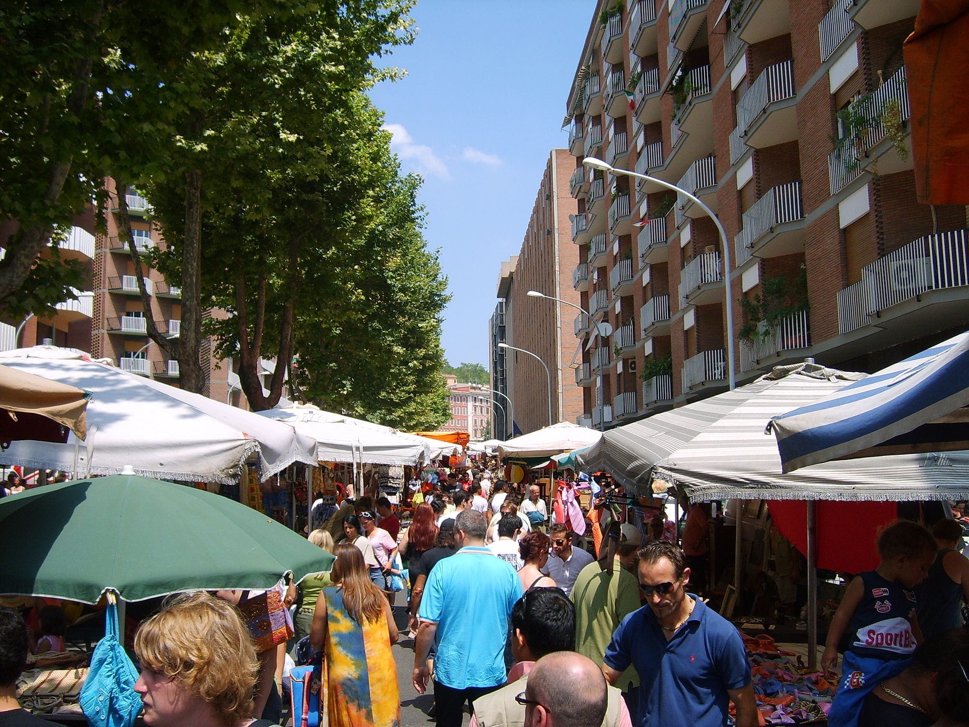 Crowds browse colorful market stalls under umbrellas at Porta Portese Flea Market, with apartment buildings and leafy trees lining a sunlit street.