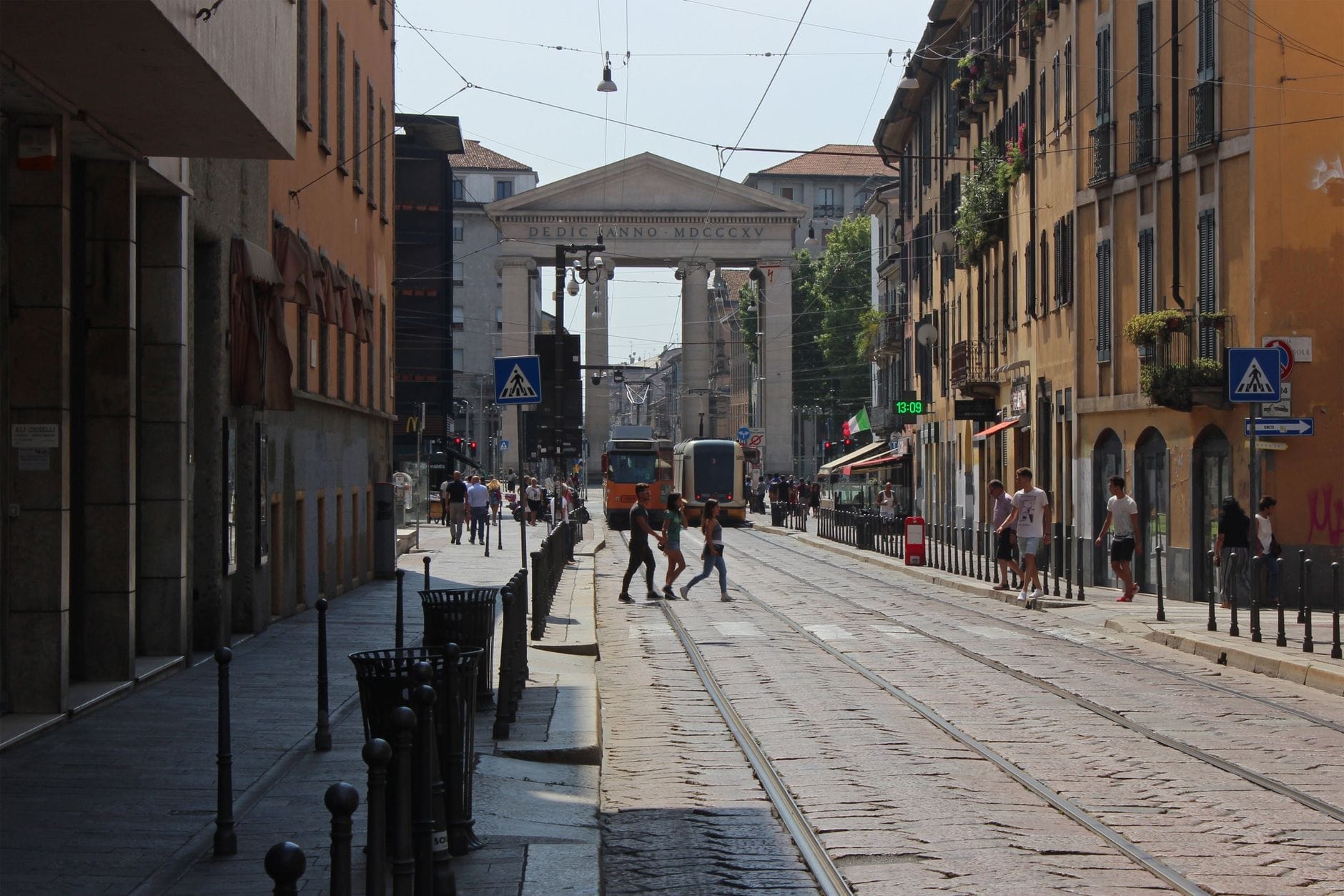 Vista della Porta Ticinese a Milano con persone che attraversano i binari del tram, la porta neoclassica sullo sfondo incorniciata da edifici storici.