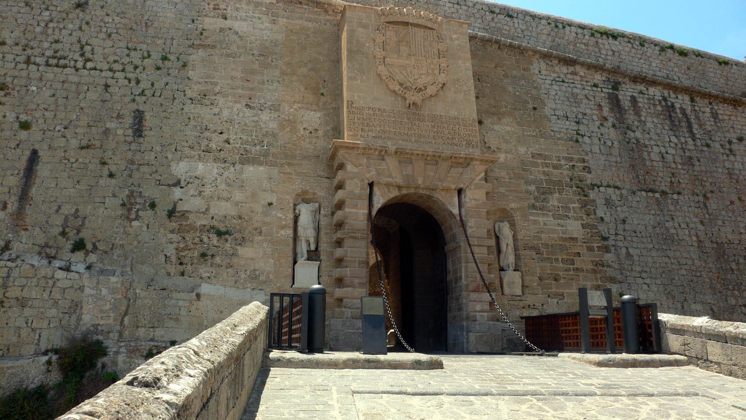 Arco d'ingresso in pietra del Portal de ses Taules a Ibiza, affiancato da statue e mura antiche sotto un cielo azzurro e soleggiato.