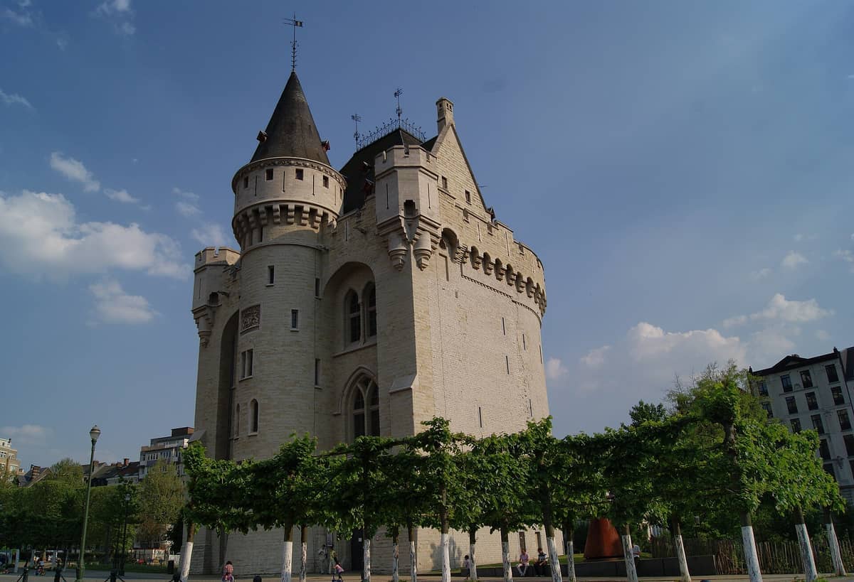 A Porte de Hal, uma guarita medieval circular de pedra com torreões, ergue-se sob um céu azul ao lado de árvores frondosas e edifícios da cidade próxima.