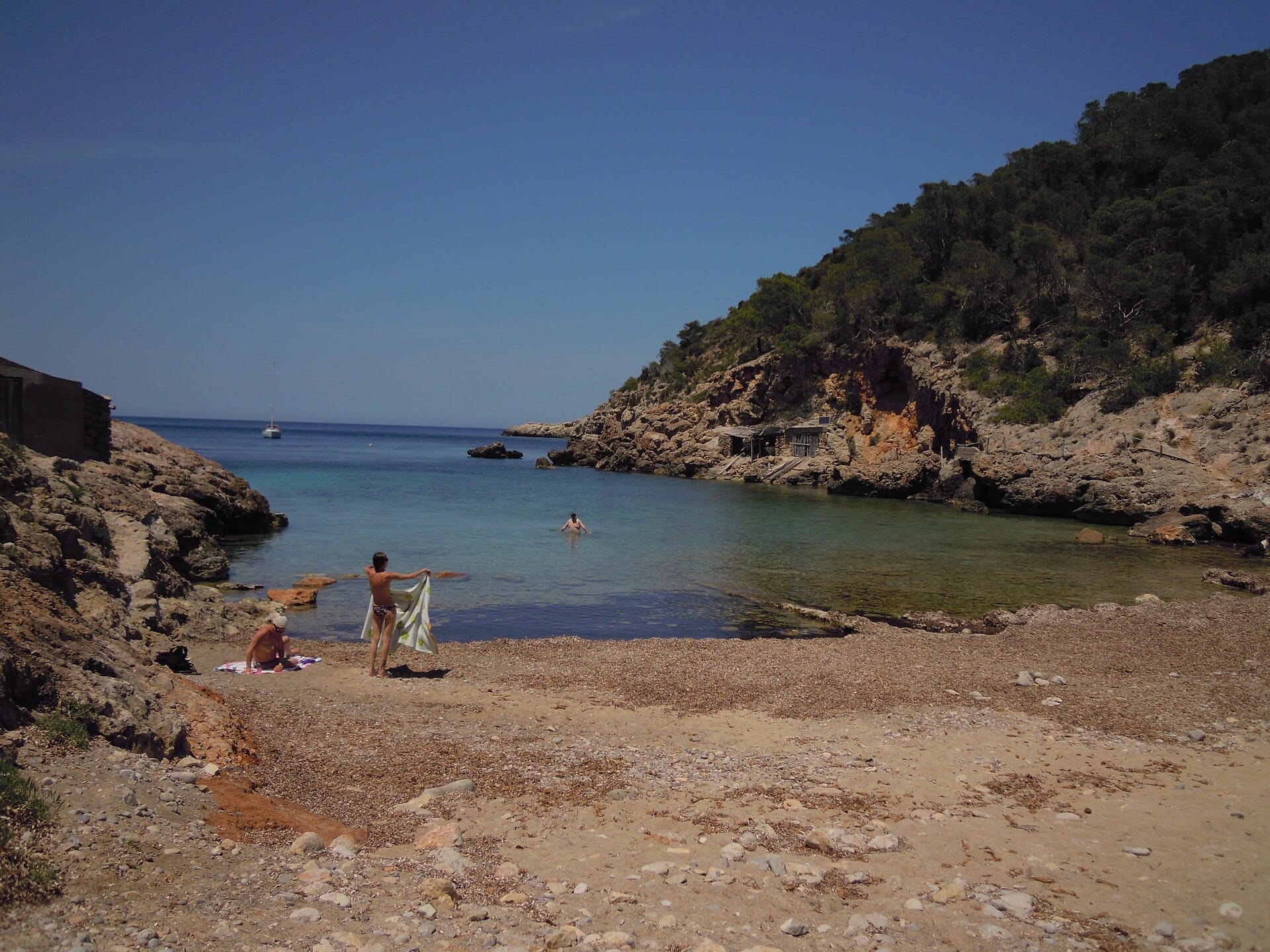 Insenatura isolata a Portinatx con acqua calma e cristallina, scogliere rocciose, rustici rimessaggi per barche e alcune persone che godono della spiaggia tranquilla.