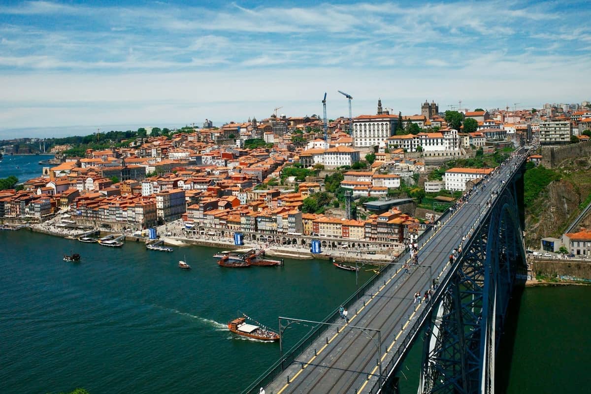 Vista aérea da Ponte Dom Luís I cruzando o rio Douro, com o colorido bairro da Ribeira do Porto empilhado ao longo da orla fluvial sob um céu azul intenso.