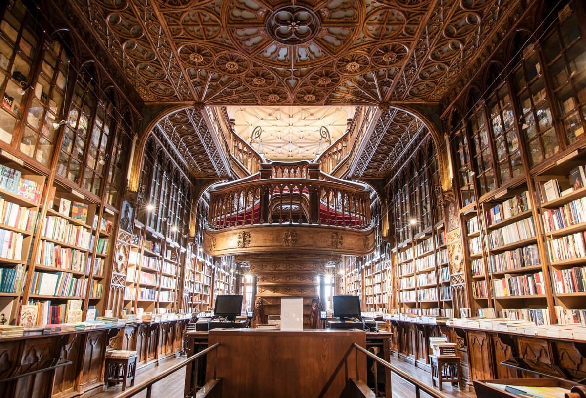 Vista ampla dos ornamentados interiores de madeira da Livraria Lello, com estantes esculpidas, uma grande escadaria e um intrincado teto decorativo.