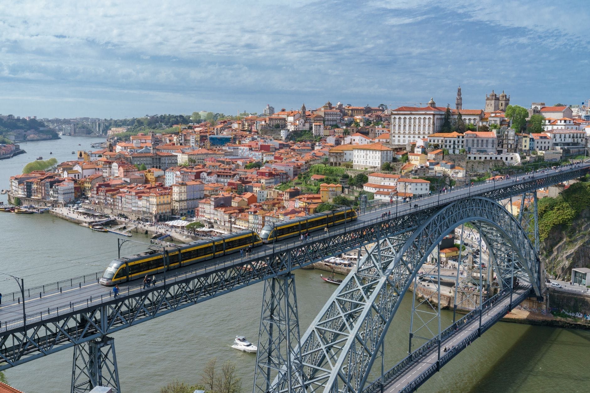 Vista panorâmica do Porto com a icônica Ponte Dom Luís I, o metro atravessando a ponte e o histórico centro da cidade às margens do Rio Douro sob um céu ensolarado.