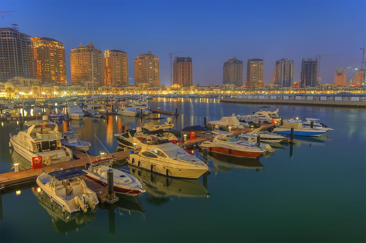 Hafenpromenade von Porto Arabia in The Pearl-Qatar am Abend, mit zahlreichen Yachten und Booten im Wasser, umgeben von hell erleuchteten Hochhäusern.