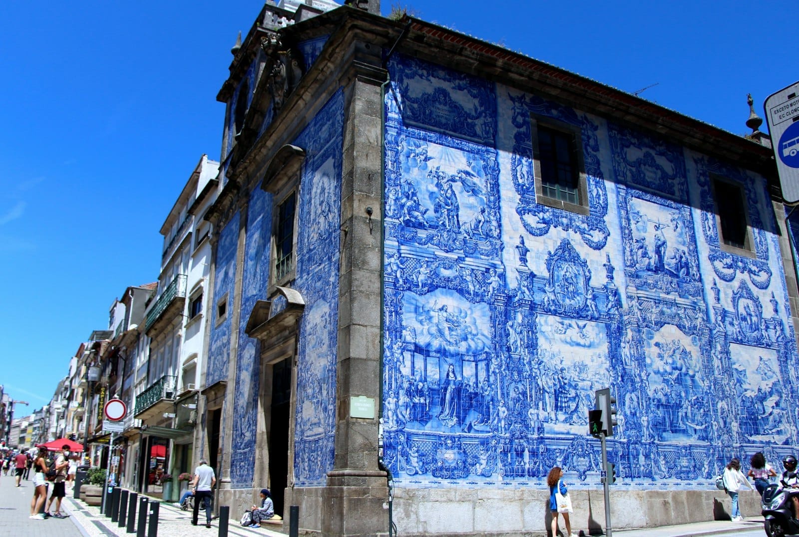 Grande painel de azulejos azuis e brancos cobrindo a lateral de uma igreja histórica no Porto, com pessoas passando em um dia ensolarado.
