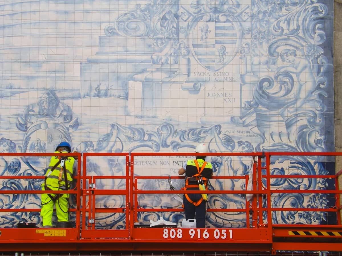 Arbeiter auf einer Hebebühne restaurieren ein großes blau-weißes Azulejo-Kachelmosaik an einer Gebäudefassade in Porto, Portugal.
