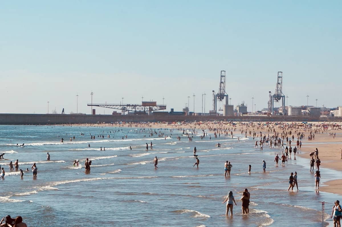 Plage urbaine animée avec des personnes qui nagent et se promènent dans les eaux peu profondes, des grues et des structures portuaires en arrière-plan.