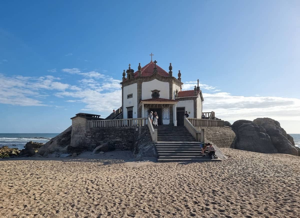 Chapelle sur la plage de Vila Nova de Gaia sous un ciel bleu, avec du sable, des rochers et l'océan Atlantique en arrière-plan.