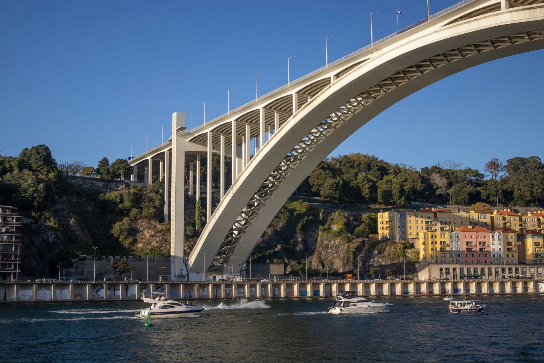 Vista ampia del Ponte dell'Arrábida che si incurva sul fiume Douro a Porto, con barche e colorati edifici sul lungofiume sotto un cielo azzurro limpido.