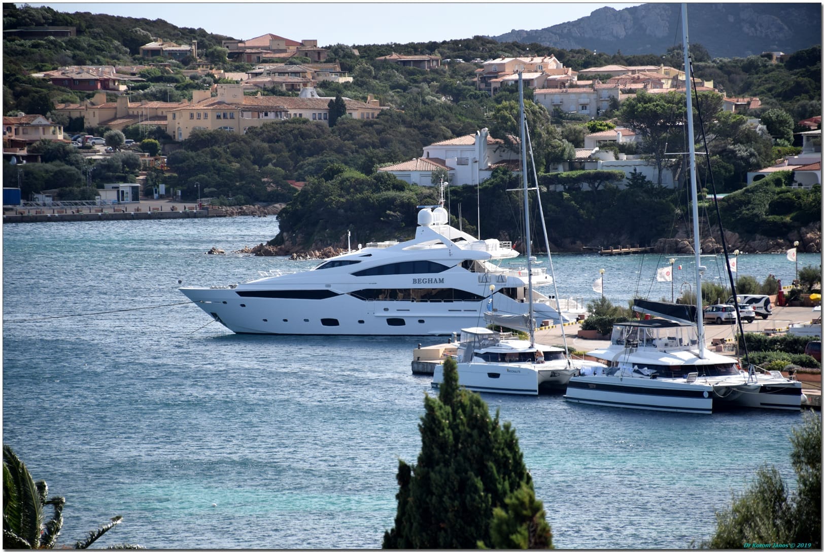 Großes weißes Superyacht und Katamarane, die in der Porto Cervo Marina anlegen, mit Hangvillen und üppigem Grün im Hintergrund.