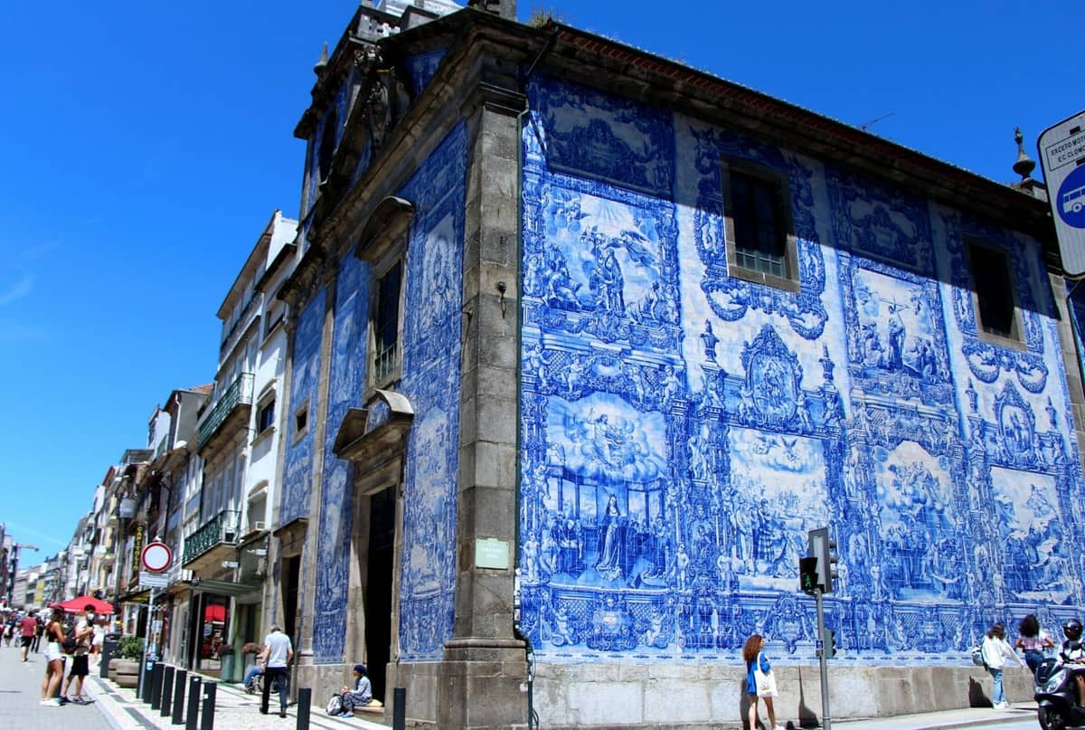 Vue en angle d'une église de Porto couverte d'azulejos bleus et blancs aux motifs complexes, avec des personnes se promenant à proximité par une journée ensoleillée.