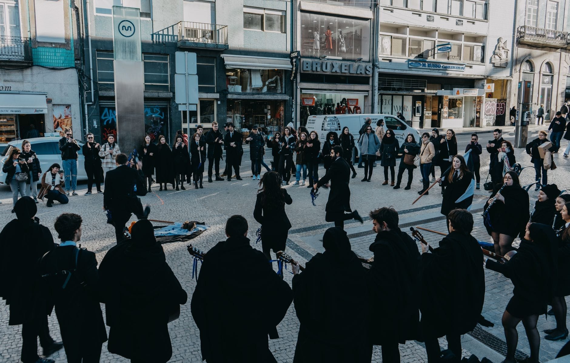 Um grupo de músicos e dançarinos se apresenta com trajes negros tradicionais numa praça central do Porto, rodeado de espectadores e edifícios da cidade.