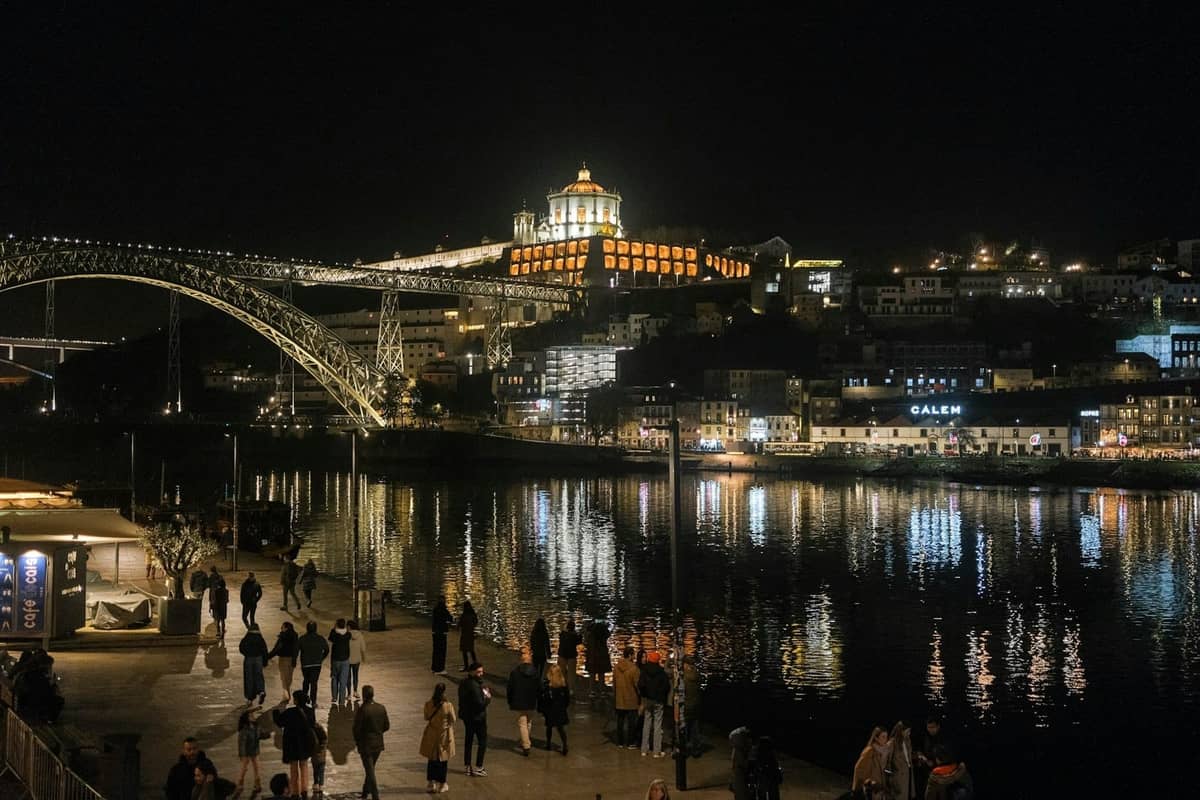 Scène nocturne le long des quais du Douro à Porto avec des promeneurs au bord de l'eau, le Pont Dom Luís I et des bâtiments illuminés en arrière-plan.