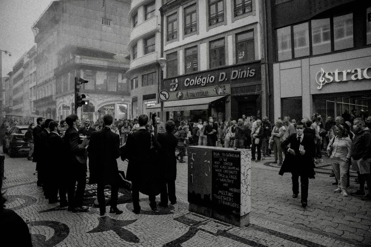 Musiciens de rue jouant en costumes noirs sur un trottoir animé de Porto, avec une foule de spectateurs et des boutiques en arrière-plan.