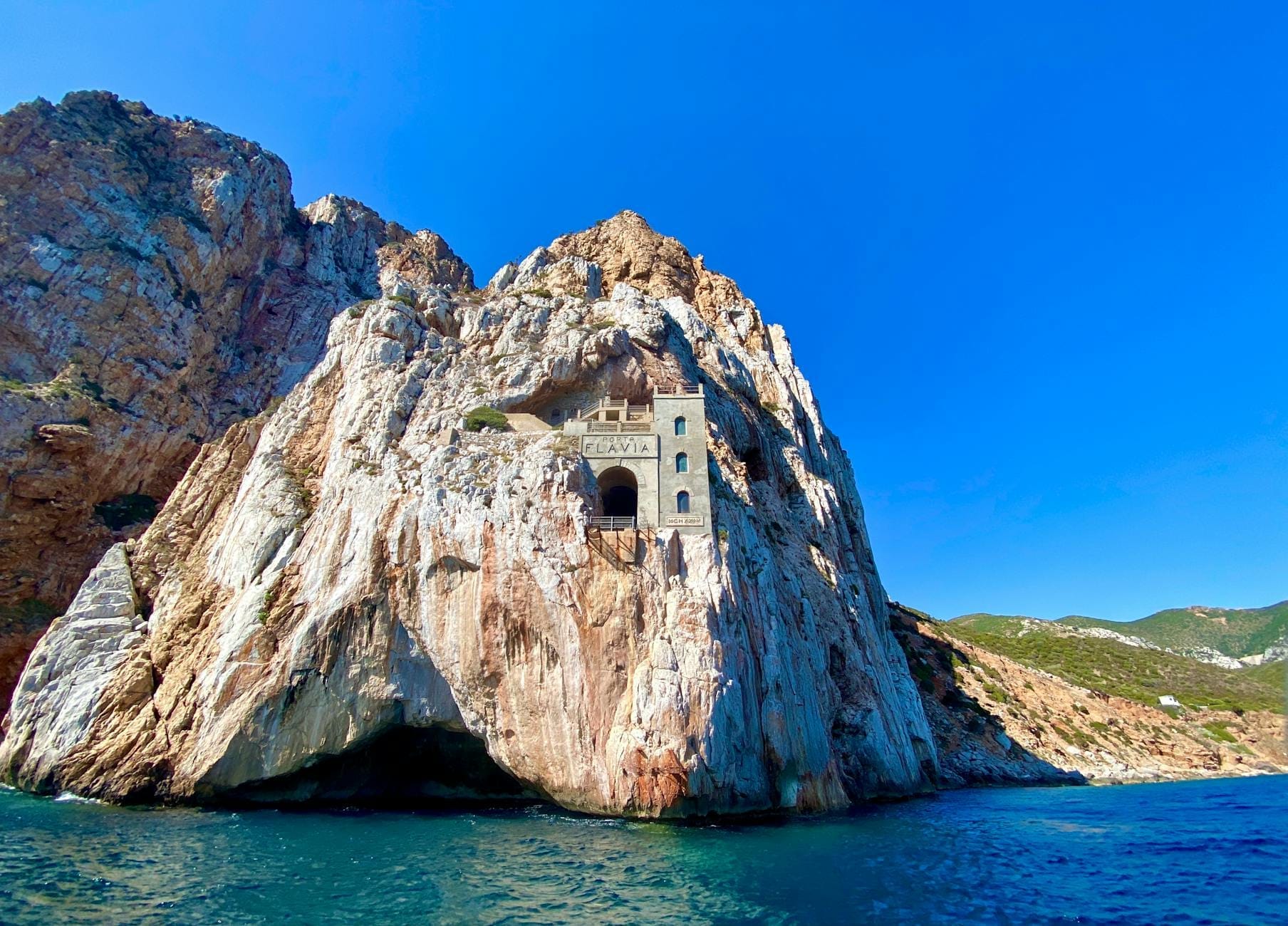 Porto Flavia, der in weiße Kalksteinklippen gehauene Bergbauhafen, dramatisch über dem blauen Meer gelegen, unter klarem Himmel, mit der sardischen Küste im Hintergrund.