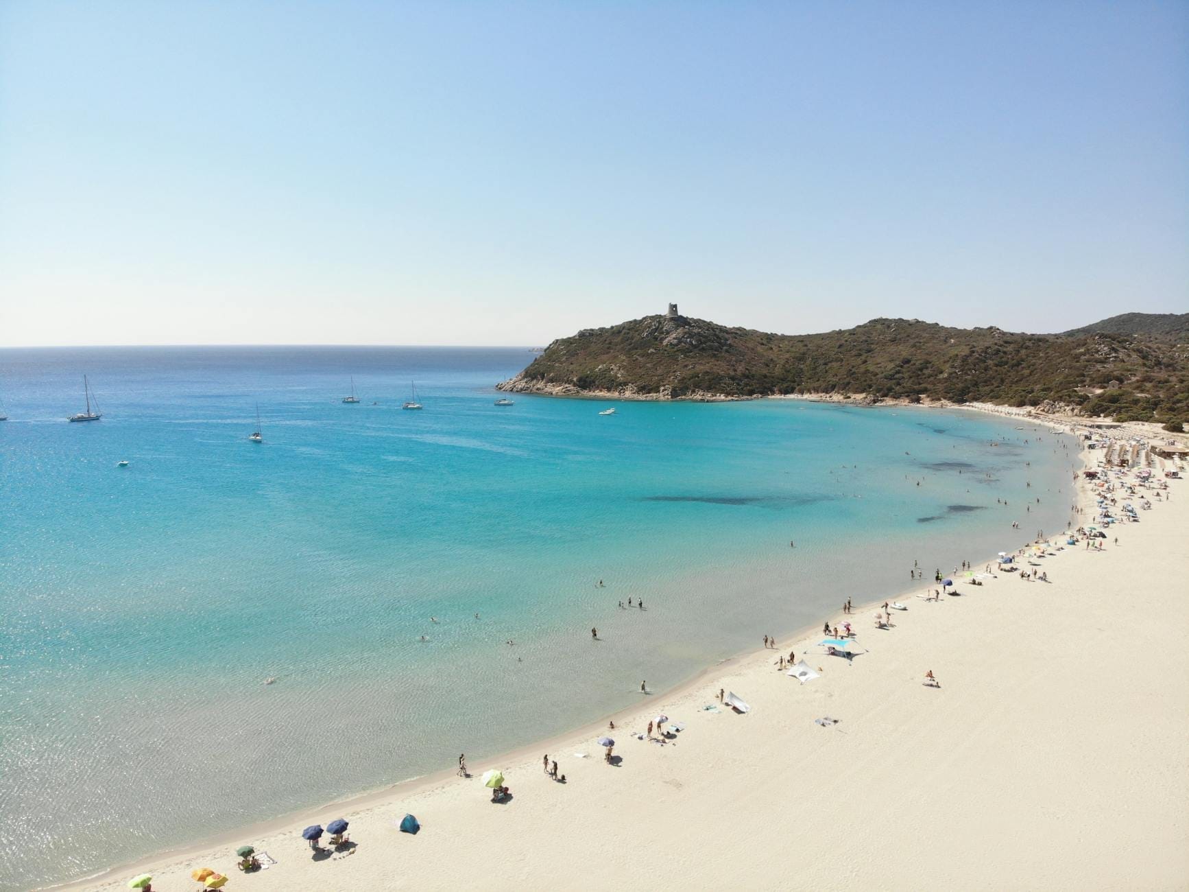 Luftaufnahme des Strandes Porto Giunco mit türkisblauem Wasser, feinem weißen Sand, Menschen beim Entspannen und dem spanischen Wachturm auf einem Hügel im Hintergrund.