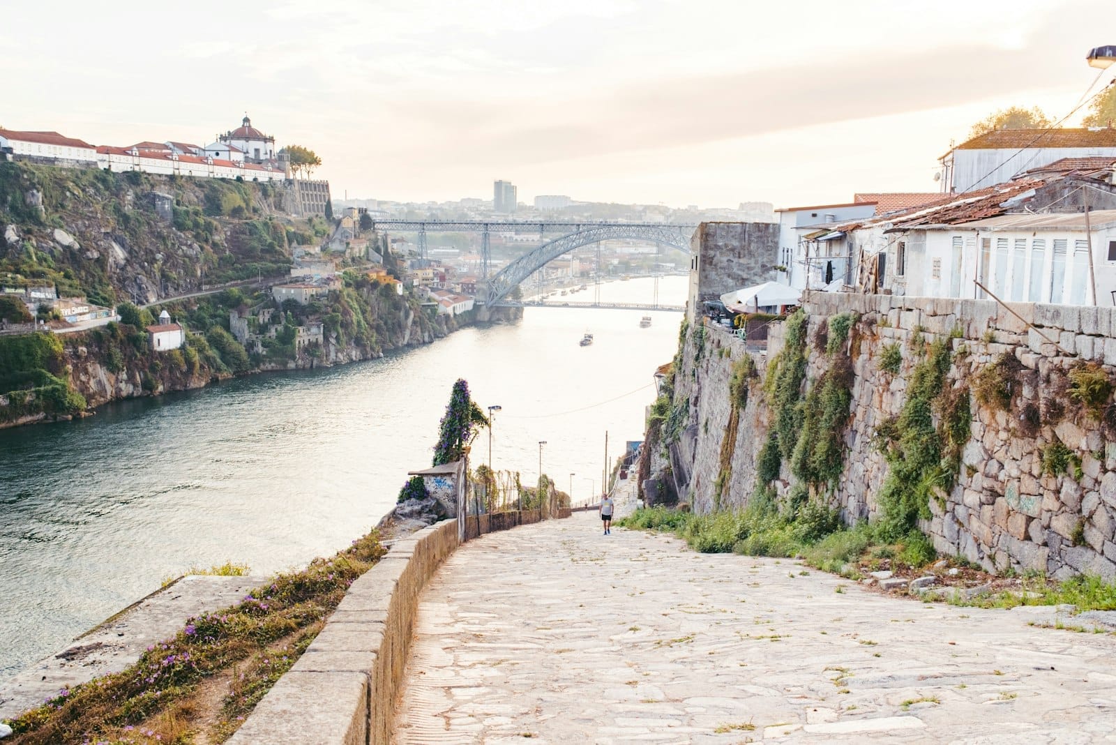 Uma calçada de pedra tranquila desce até o Rio Douro com a icônica ponte de ferro do Porto ao fundo e prédios iluminados pelo sol nas duas margens.