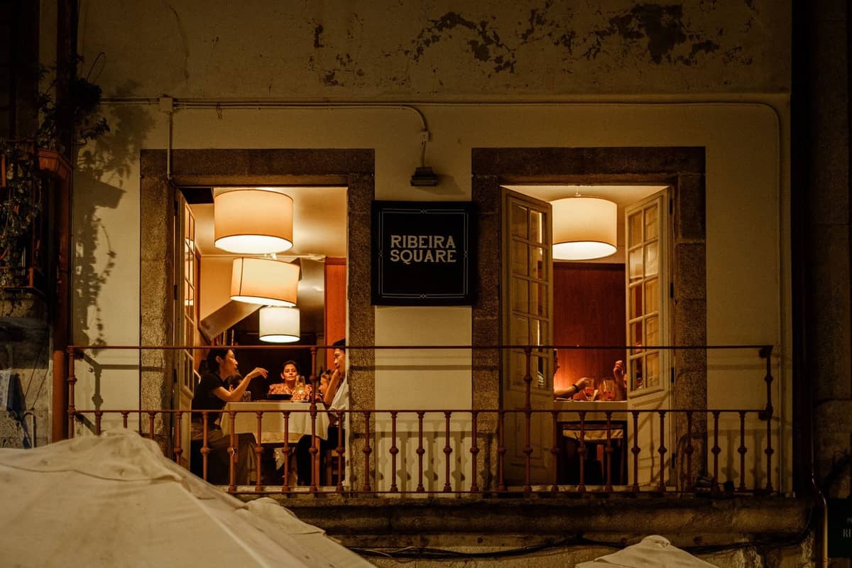 Vista con luce calda di un ristorante in Piazza Ribeira a Porto di notte, con i commensali visibili attraverso le finestre aperte.