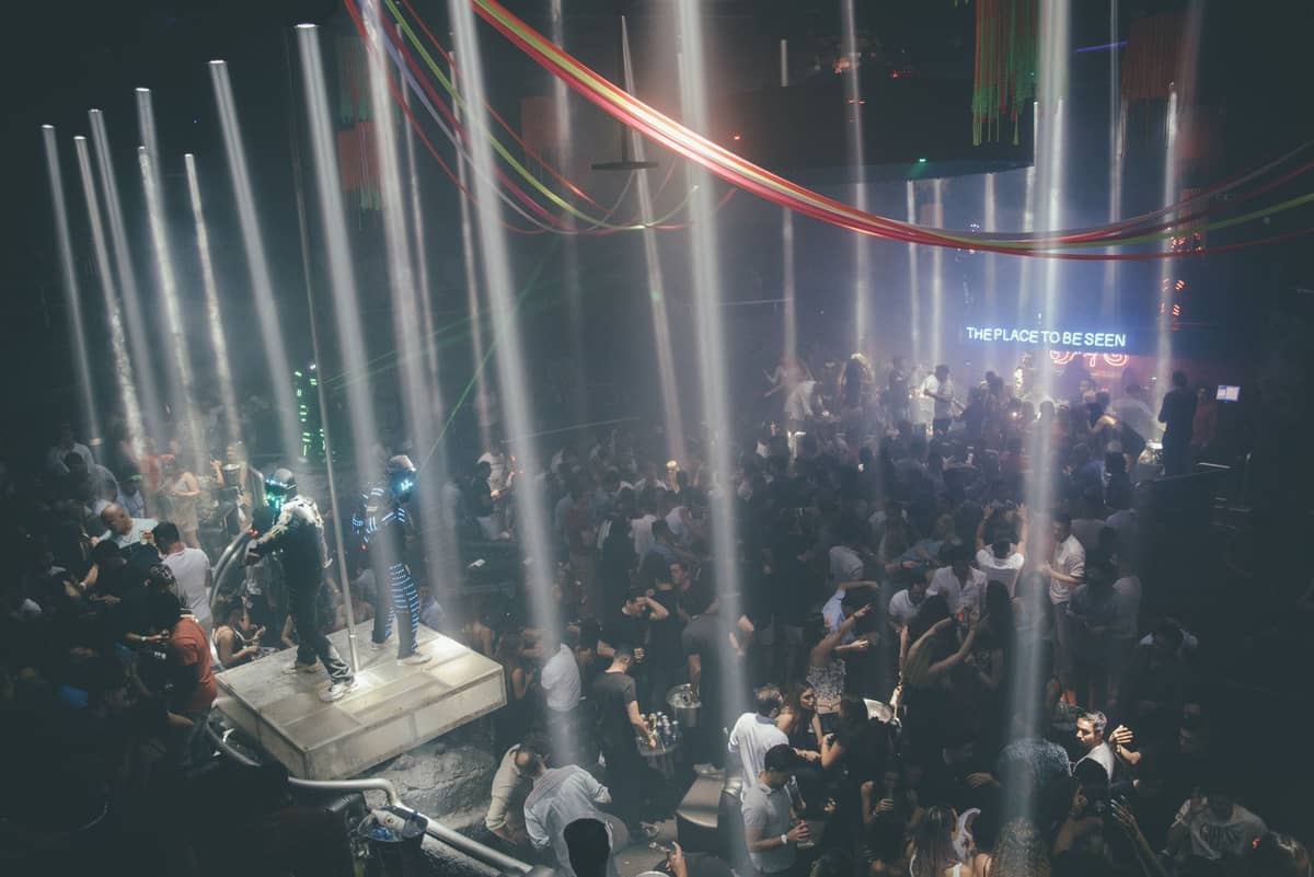 Bir platformda dansçılar ve arka planda parlak bir ekranla ışık huzmelerinin altında büyük bir gece kulübü kalabalığı.