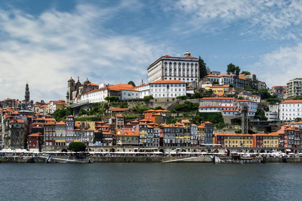 Vista panoramica dello storico lungofiume di Porto con colorati edifici antichi, il fiume Douro e l'architettura collinare sotto un cielo luminoso.