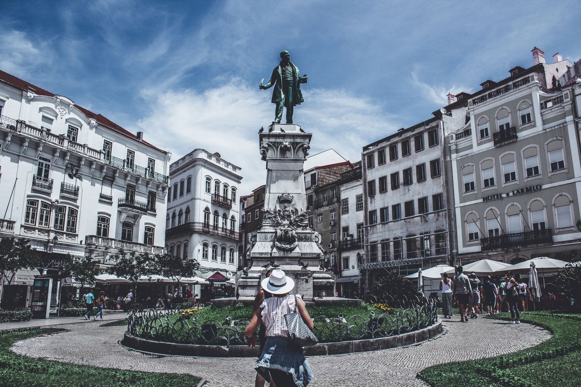 Pessoas caminham e relaxam perto da estátua de Dom Pedro IV em uma praça animada no Porto, cercada por clássicos edifícios brancos e um céu vibrante.