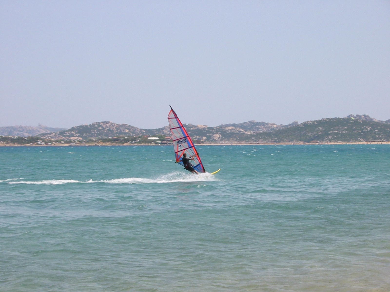 Ein Windsurfer gleitet über das türkisfarbene Wasser von Porto Pollo, mit Sardiniens felsiger Küste unter einem klaren blauen Himmel.