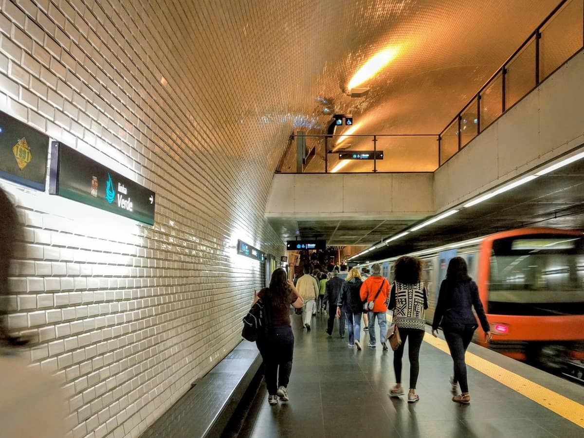 Fahrgäste gehen auf einem gut beleuchteten U-Bahnsteig in Porto entlang, während ein U-Bahnzug einfährt und Stationsschilder an der gefliesten Wand sichtbar sind.