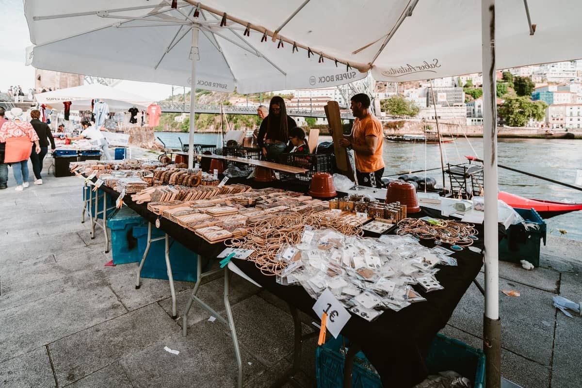 Marktstand im Freien am Fluss in Porto mit lokalen Kunsthandwerken und Lebensmitteln unter weißen Sonnenschirmen.