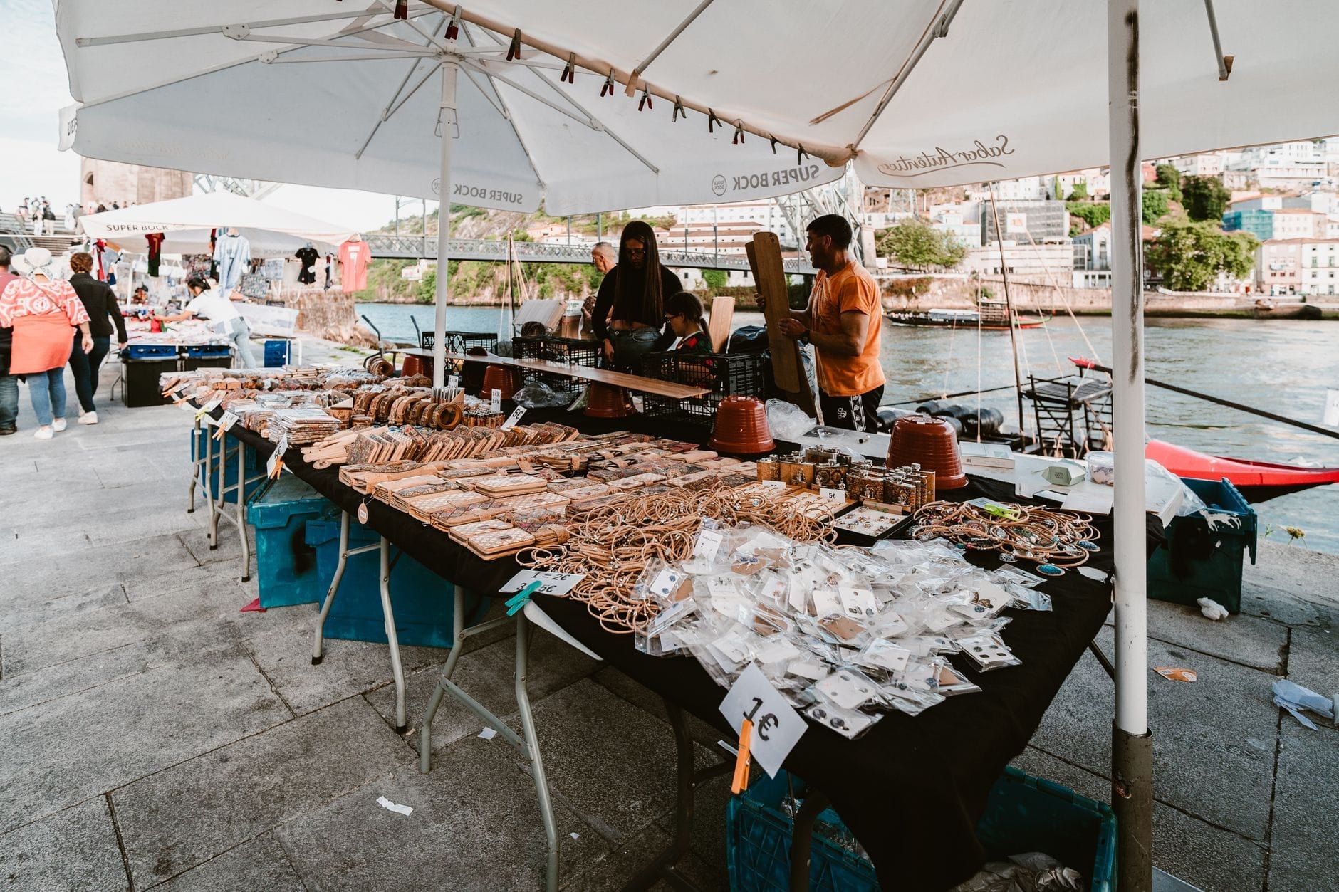 Mercado ao ar livre à beira do rio no Porto, com barracas vendendo artesanato e comida sob guarda-sóis brancos, pessoas passeando e o rio ao fundo.