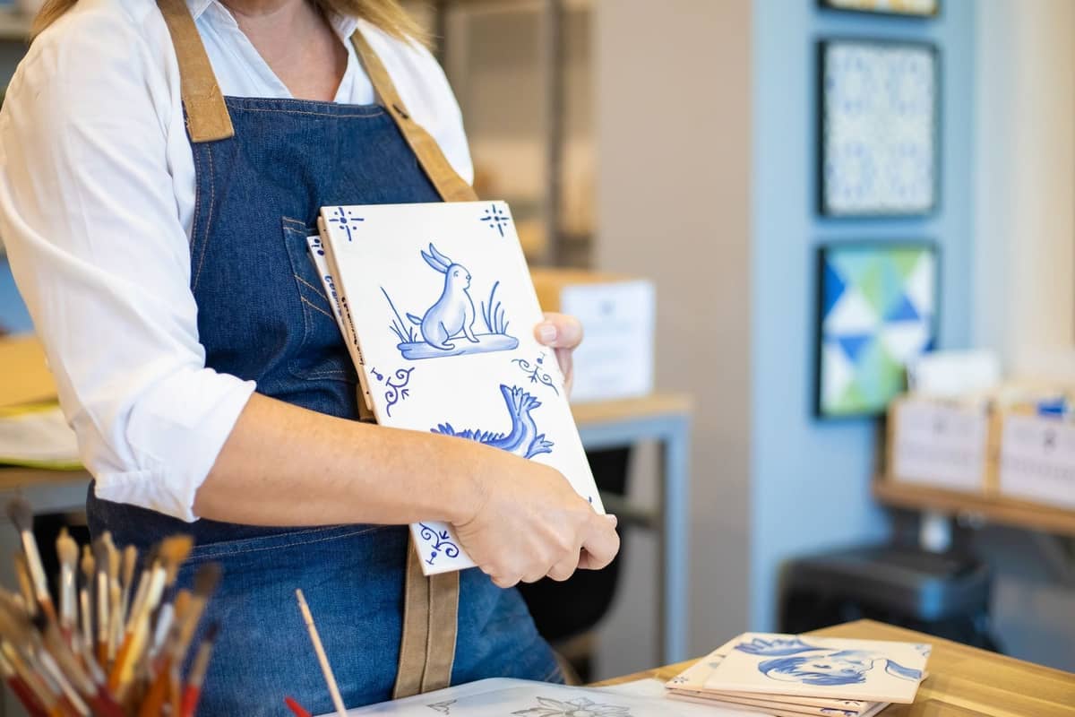 Person in einer Schürze hält handbemalte portugiesische Keramikfliesen in Blau und Weiß, umgeben von Malwerkzeugen in einem Studio oder Geschäft.
