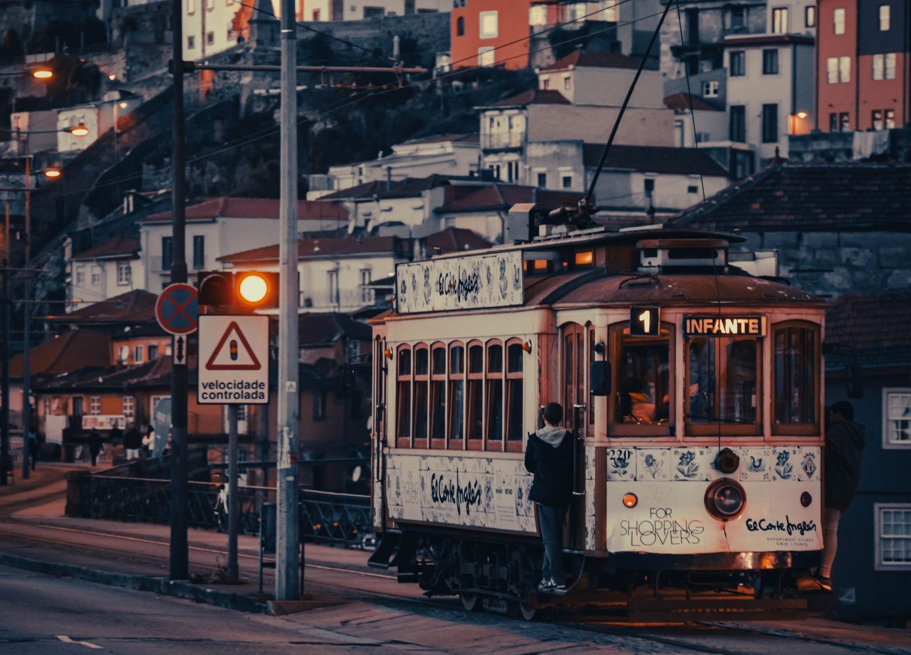 Un tram d'epoca con la scritta Infante percorre la storica Linea 1 di Porto lungo il fiume Douro al tramonto, con le colorate case sulla collina sullo sfondo.