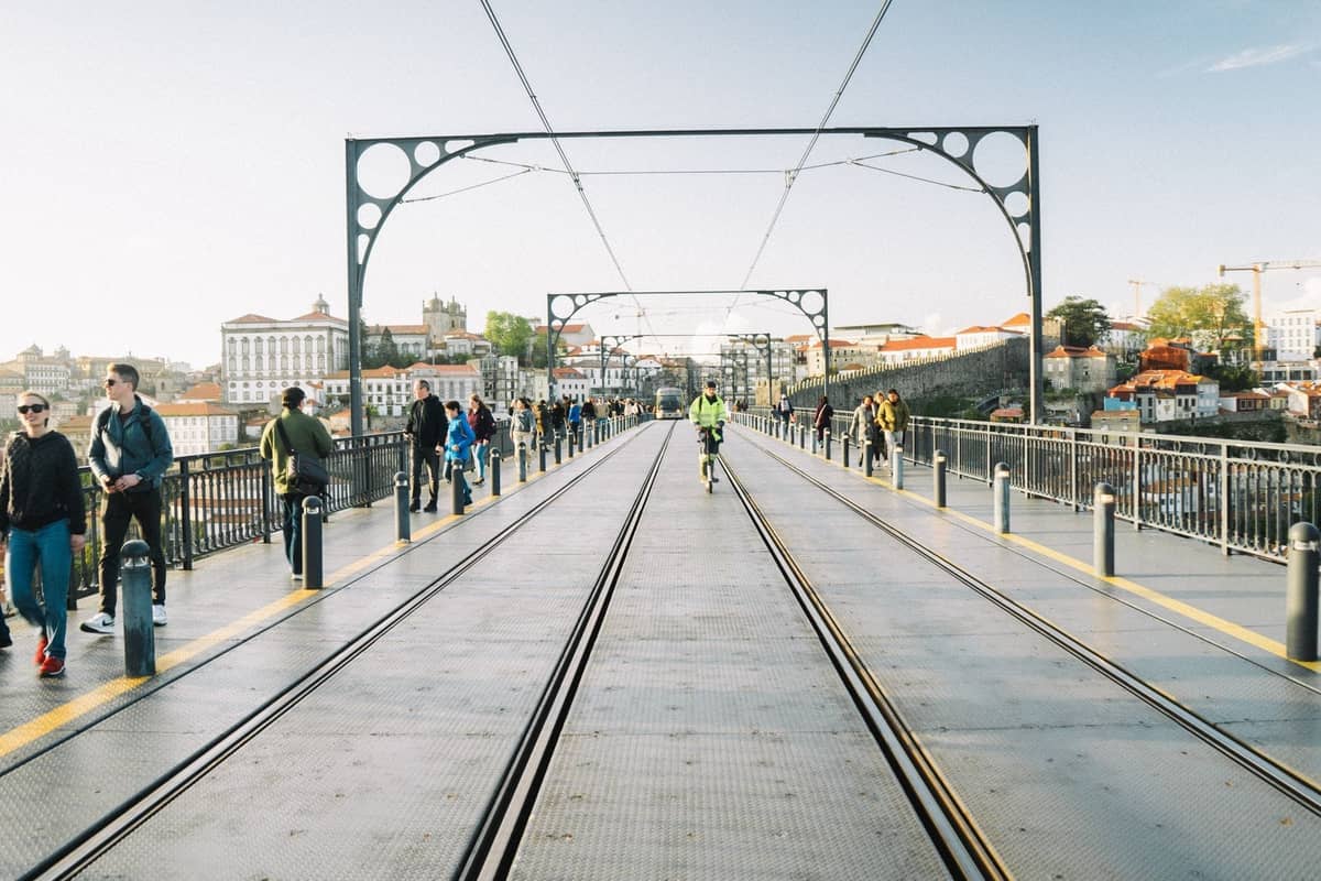 Turisti e abitanti del posto che camminano sul iconico Ponte Dom Luís I a Porto, con vedute della città e un cielo sereno.