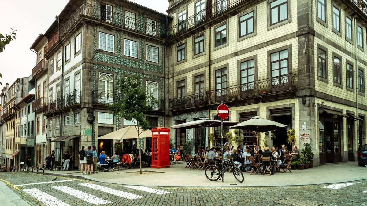Vista en esquina de un animado barrio de Porto con edificios de azulejos, terrazas de café, personas comiendo y una cabina telefónica roja de estilo británico en una calle empedrada.