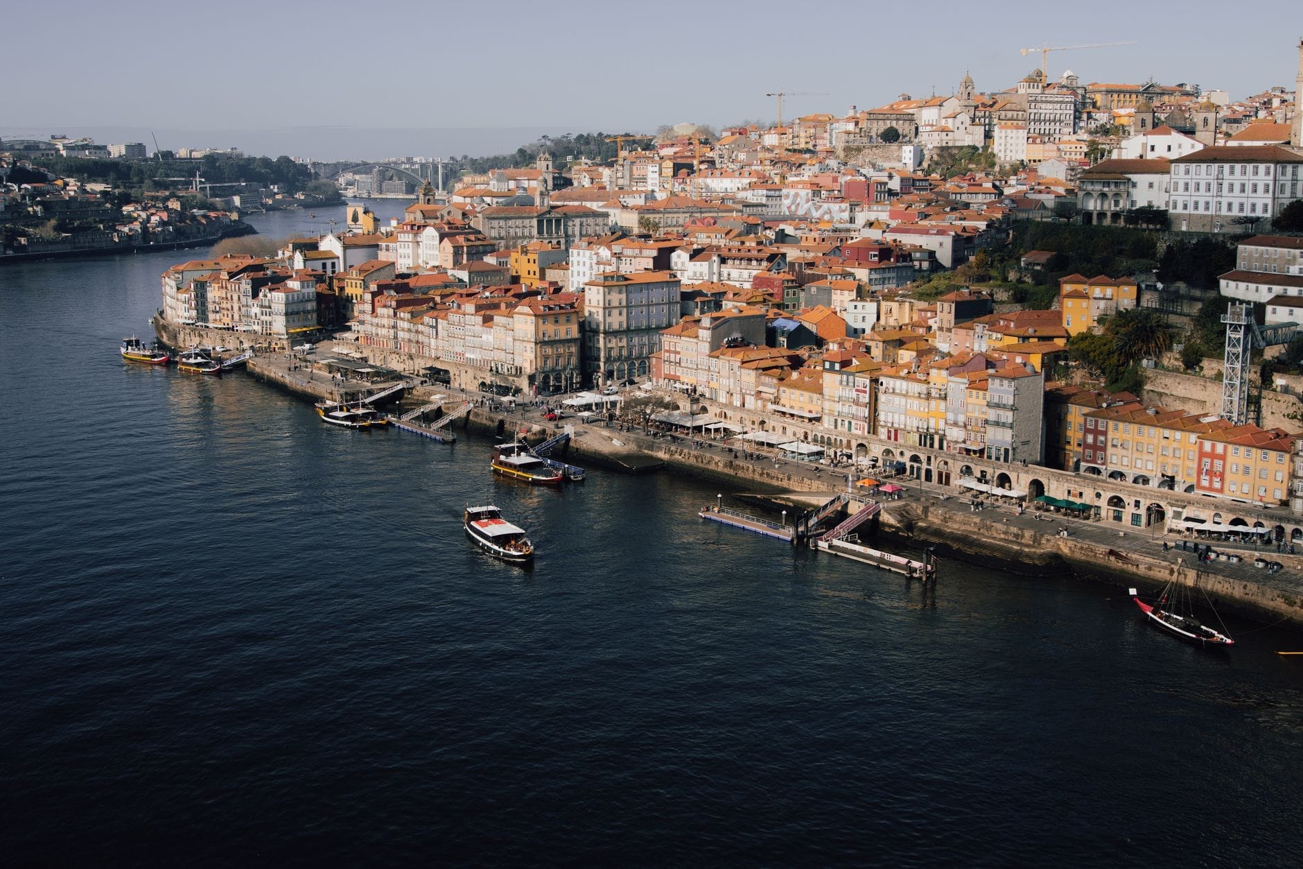 Vista panorâmica das coloridas construções históricas à beira-rio de Porto e dos barcos alinhados no rio Douro, com ruas inclinadas subindo a partir da água sob a luz clara do dia.