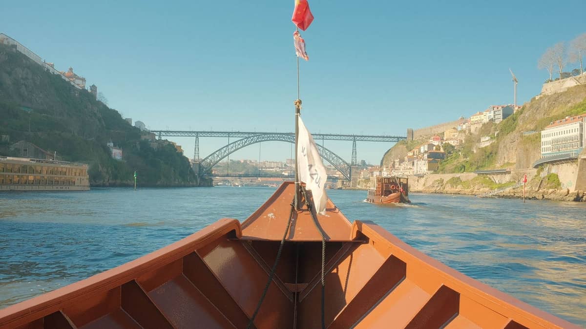 Blick von einem Boot auf dem Fluss Douro in Porto, in Richtung einer ikonischen Brücke und malerischer Stadthänge unter blauem Himmel.