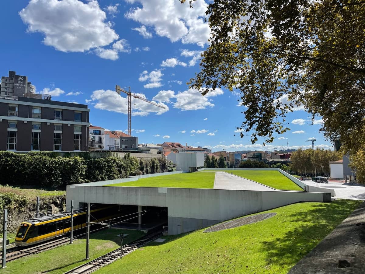 Außenansicht eines Metro do Porto-Zuges, der in einen modernen Bahnhof einfährt, umgeben von Grünflächen und Stadtgebäuden unter blauem Himmel.