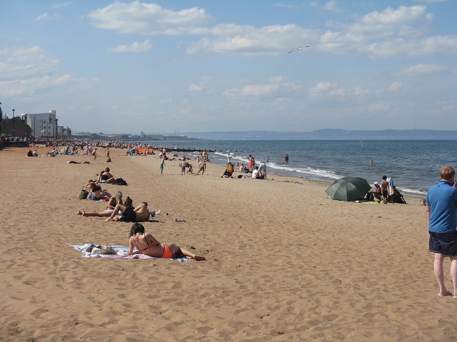 Menschen, die sich auf dem Sandstreifen von Portobello Beach entspannen, mit dem blauen Meer, der viktorianischen Promenade und entfernten Gebäuden unter einem teilweise bewölkten Himmel.