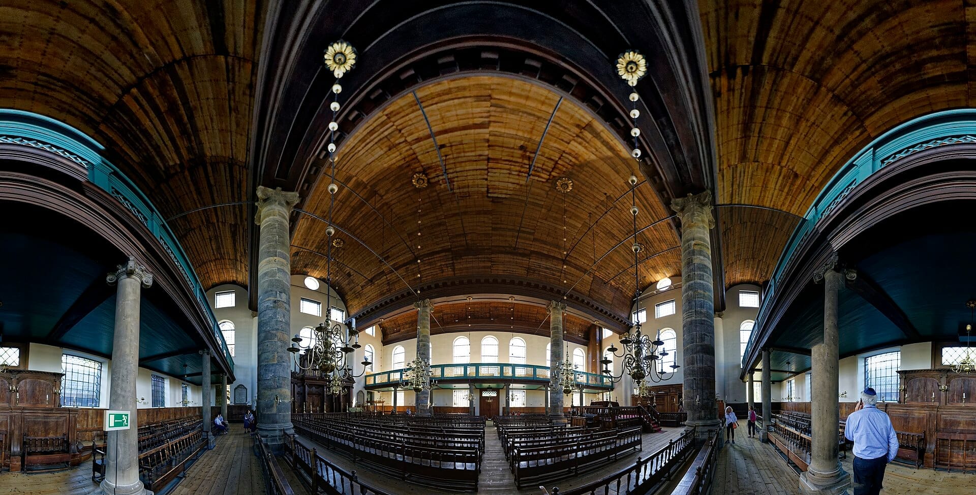 Vista gran angular del interior de la Sinagoga Portuguesa de Ámsterdam, con techo abovedado de madera, columnas de piedra, candelabros y bancos de madera con luz natural.