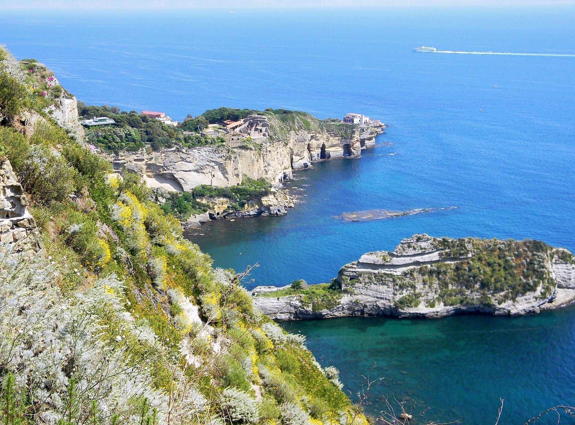 Vista spettacolare sulla scogliera del Parco Virgiliano con il Golfo di Napoli sullo sfondo, il mare di un blu intenso, la vegetazione rigogliosa e le formazioni rocciose frastagliate in primo piano.