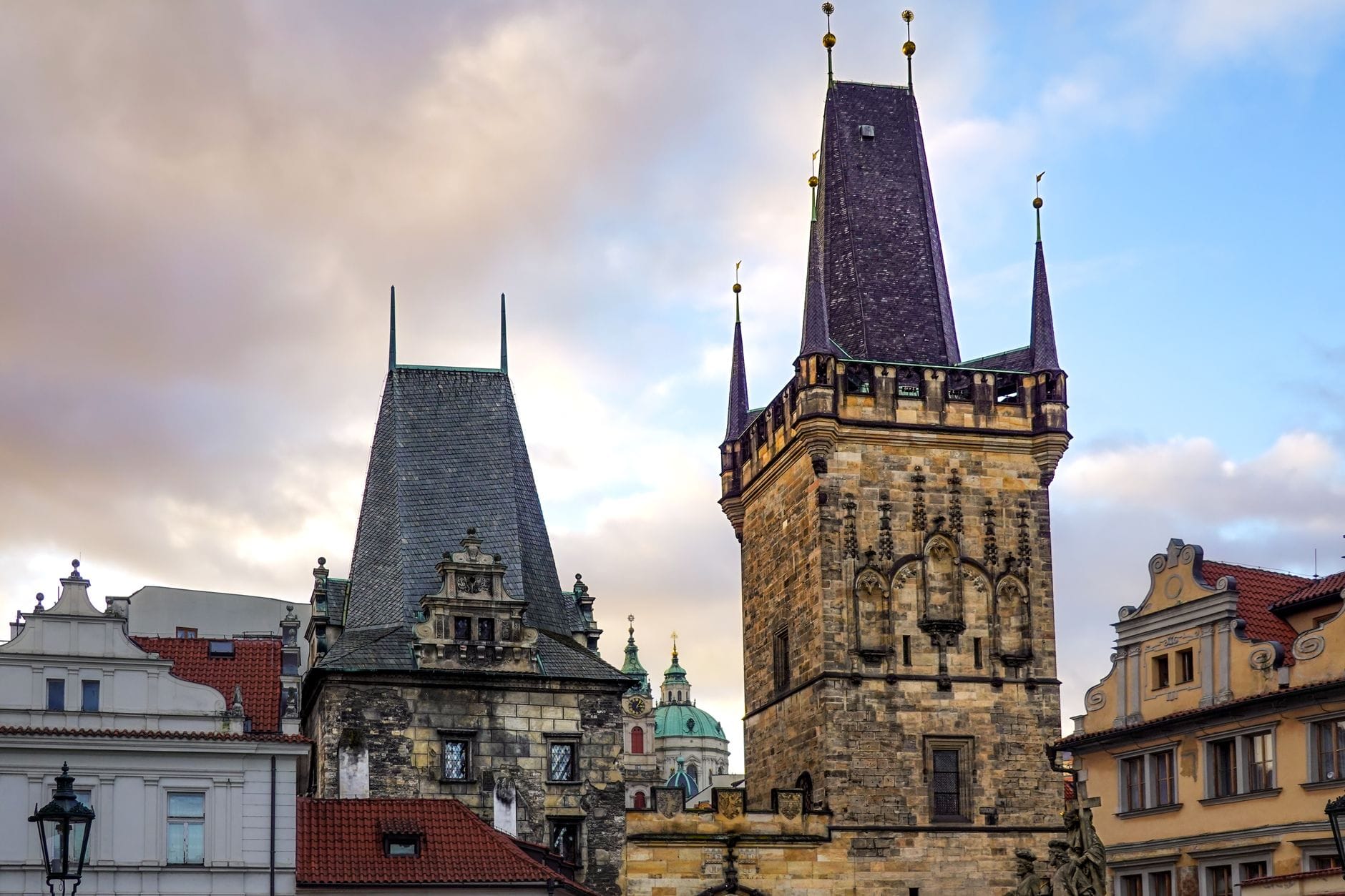 Der Pulverturm in Prag mit seiner hohen, reich verzierten gotischen Architektur, der im Abendlicht zwischen historischen Nachbargebäuden unter einem dramatischen Himmel prominent aufragt.