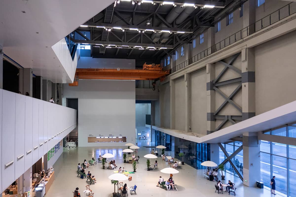 Innenansicht des Power Station of Art in Shanghai: eine helle, weitläufige Industriehalle mit hohen Decken, großen Fenstern, Menschen an Tischen und modernen Architekturelementen.