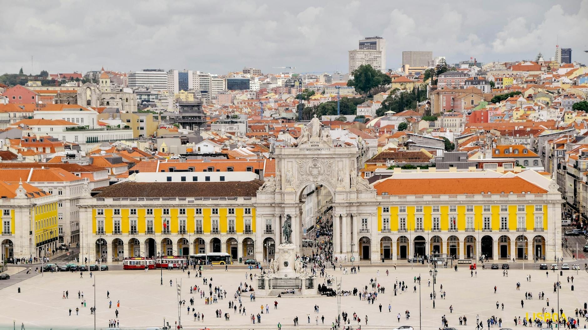 Una vista aerea grandangolare della Praça do Comércio con la sua imponente piazza, i caratteristici edifici gialli, l'arco centrale e il panorama di Lisbona sullo sfondo.